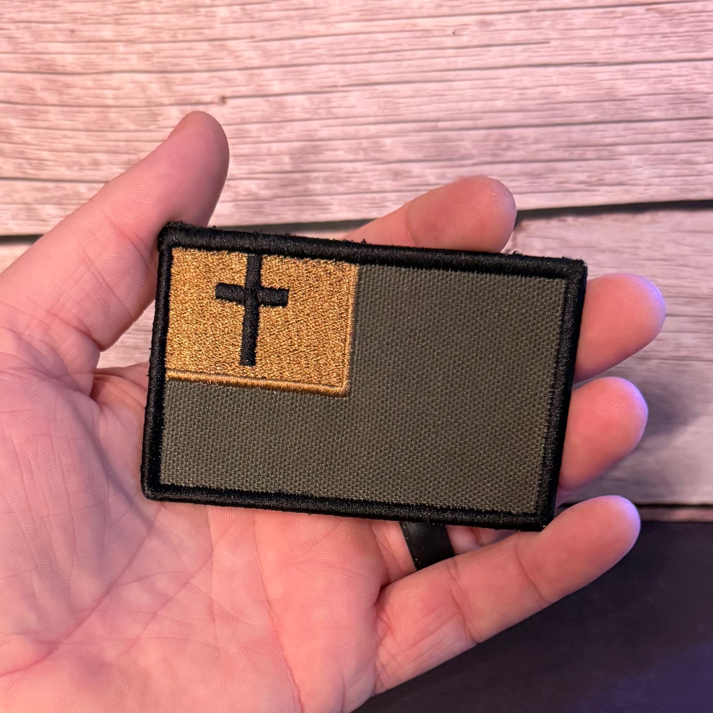 A rectangular embroidered patch held in a hand, featuring an olive drab green background with a tan square in the upper left containing a black cross, set against a light wood plank background.