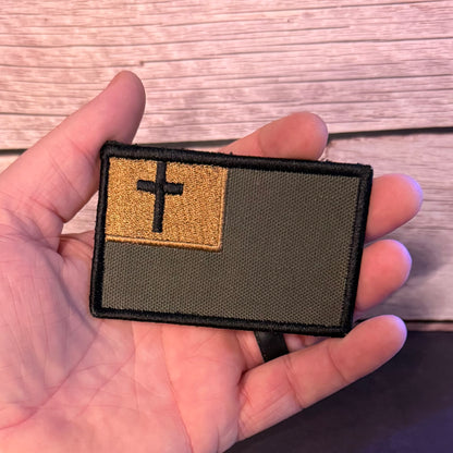 A rectangular embroidered patch held in a hand, featuring an olive drab green background with a tan square in the upper left containing a black cross, set against a light wood plank background.