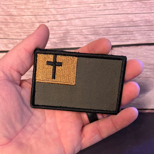 A rectangular embroidered patch held in a hand, featuring an olive drab green background with a tan square in the upper left containing a black cross, set against a light wood plank background.
