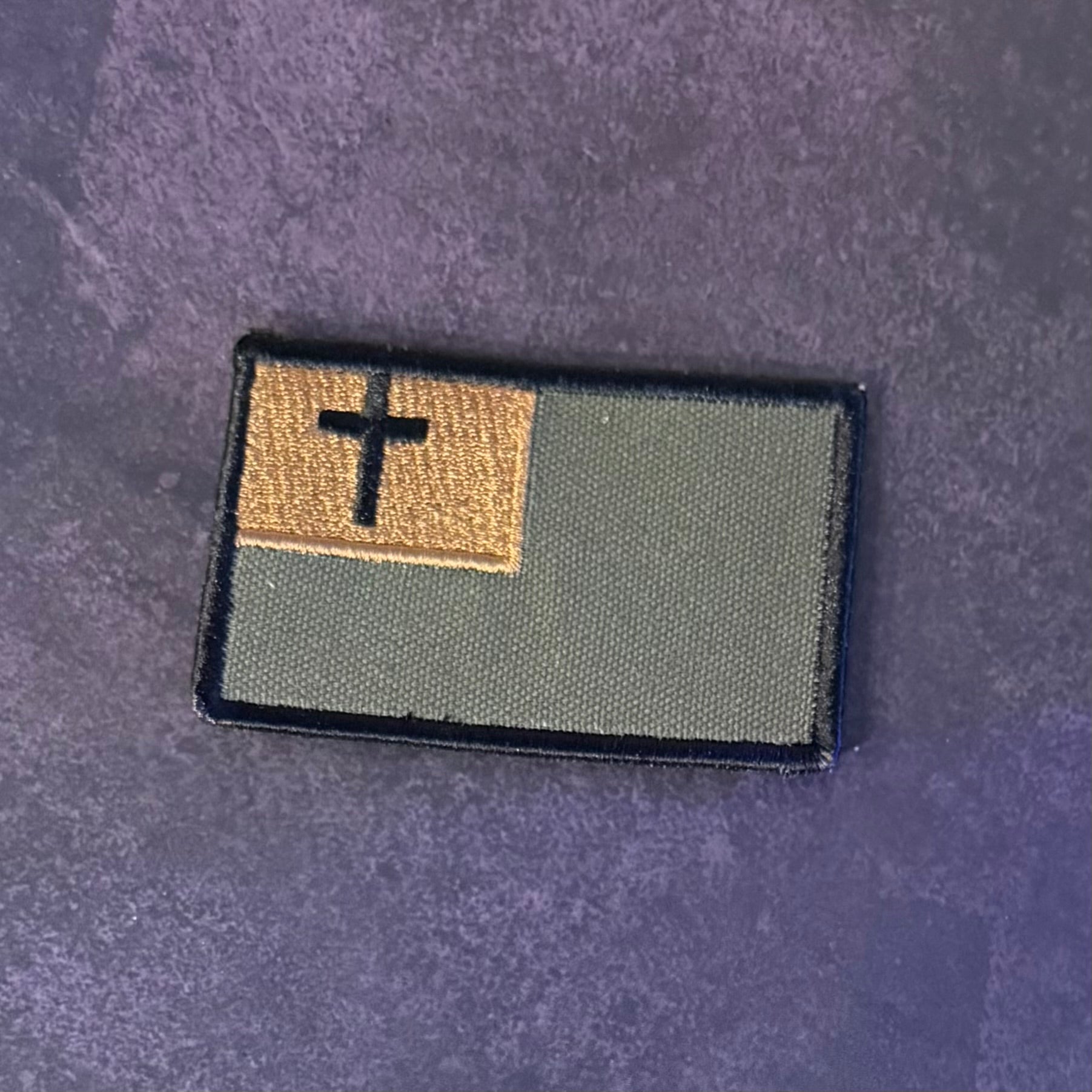 An embroidered rectangular patch featuring a black cross on a tan square in the upper left, with an olive green field and black border, placed on a textured dark purple background.