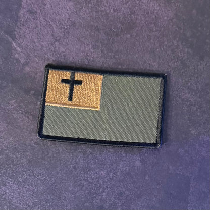 An embroidered rectangular patch featuring a black cross on a tan square in the upper left, with an olive green field and black border, placed on a textured dark purple background.
