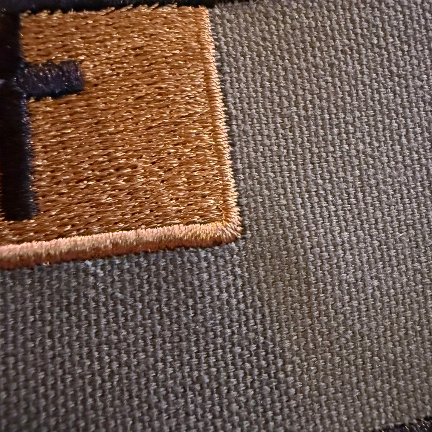Close-up of a woven fabric surface with a tan embroidered square patch and a partial black embroidered letter on the left, showing the texture of the material.