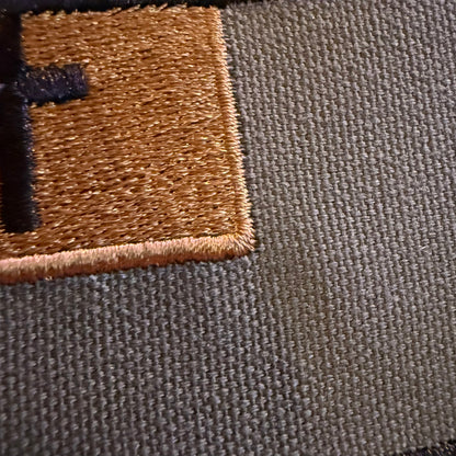 Close-up of a woven fabric surface with a tan embroidered square patch and a partial black embroidered letter on the left, showing the texture of the material.