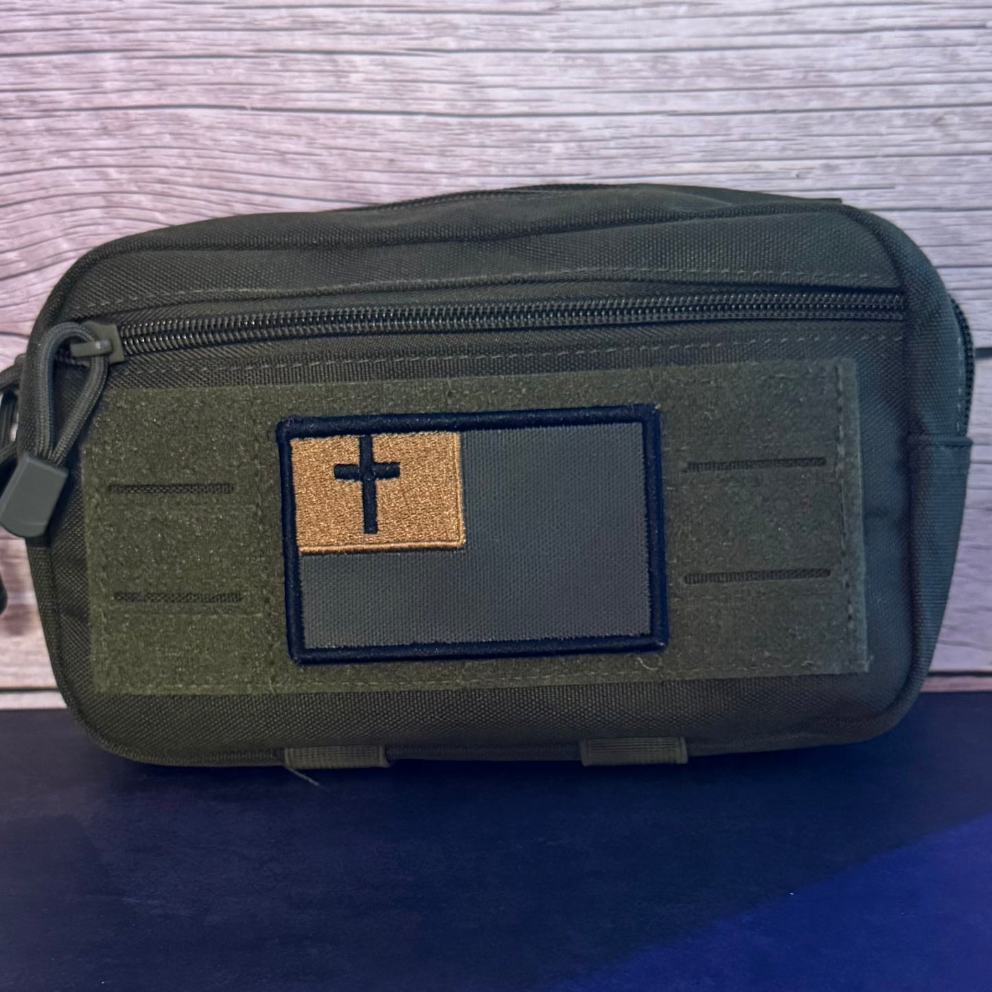 An olive green tactical pouch with a front zippered pocket and a rectangular patch featuring a black cross on a tan background, set against a wood-grain backdrop.