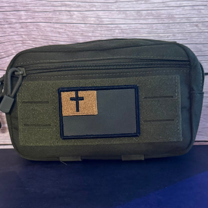 An olive green tactical pouch with a front zippered pocket and a rectangular patch featuring a black cross on a tan background, set against a wood-grain backdrop.