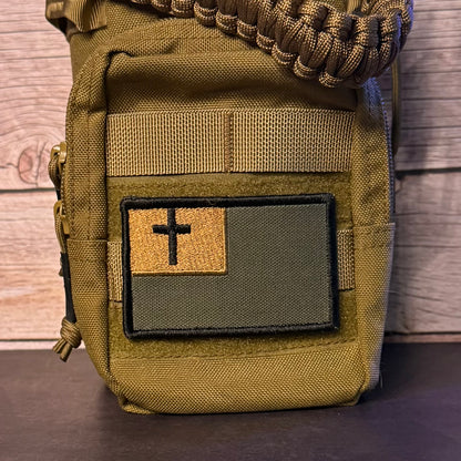 An olive drab tactical pouch with a front patch featuring a black cross on a split tan and dark green background, shown against a wood plank backdrop.