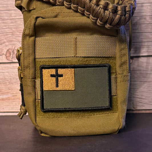 An olive drab tactical pouch with a front patch featuring a black cross on a split tan and dark green background, shown against a wood plank backdrop.