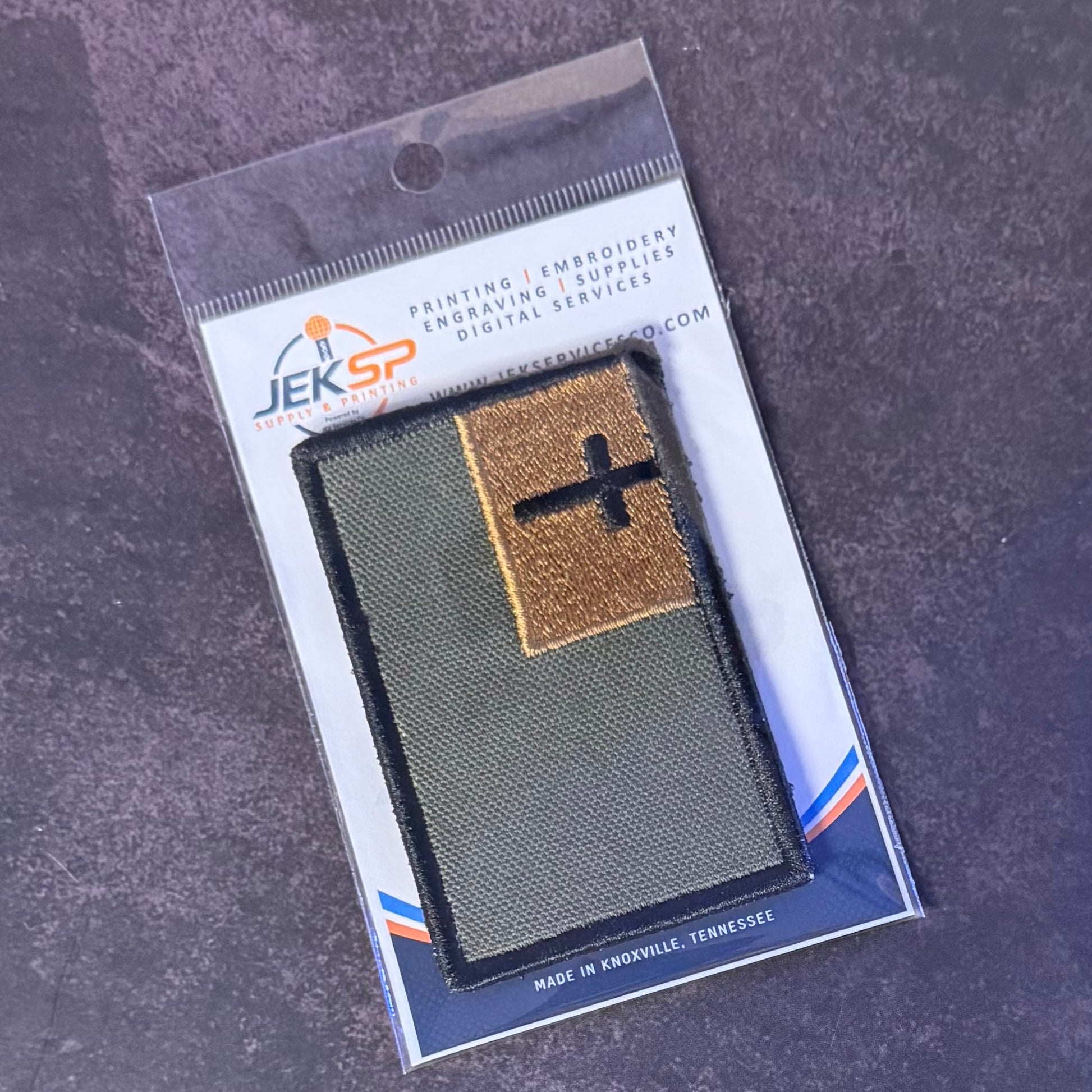 An embroidered military-style patch in its retail blister packaging, photographed on a dark textured surface. The patch is olive green with a tan square section containing a black cross, and the backing card shows the brand JEKSP.