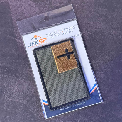 An embroidered military-style patch in its retail blister packaging, photographed on a dark textured surface. The patch is olive green with a tan square section containing a black cross, and the backing card shows the brand JEKSP.
