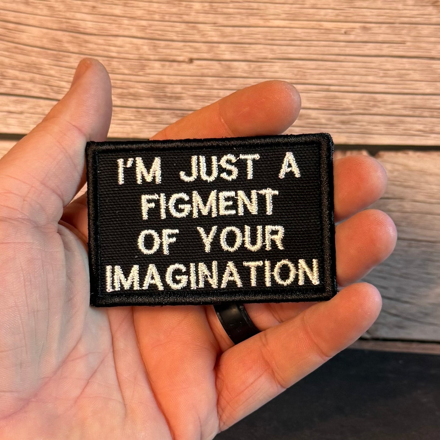 A hand holding a rectangular black embroidered patch with white text that reads, 'I'M JUST A FIGMENT OF YOUR IMAGINATION' against a wood plank background.