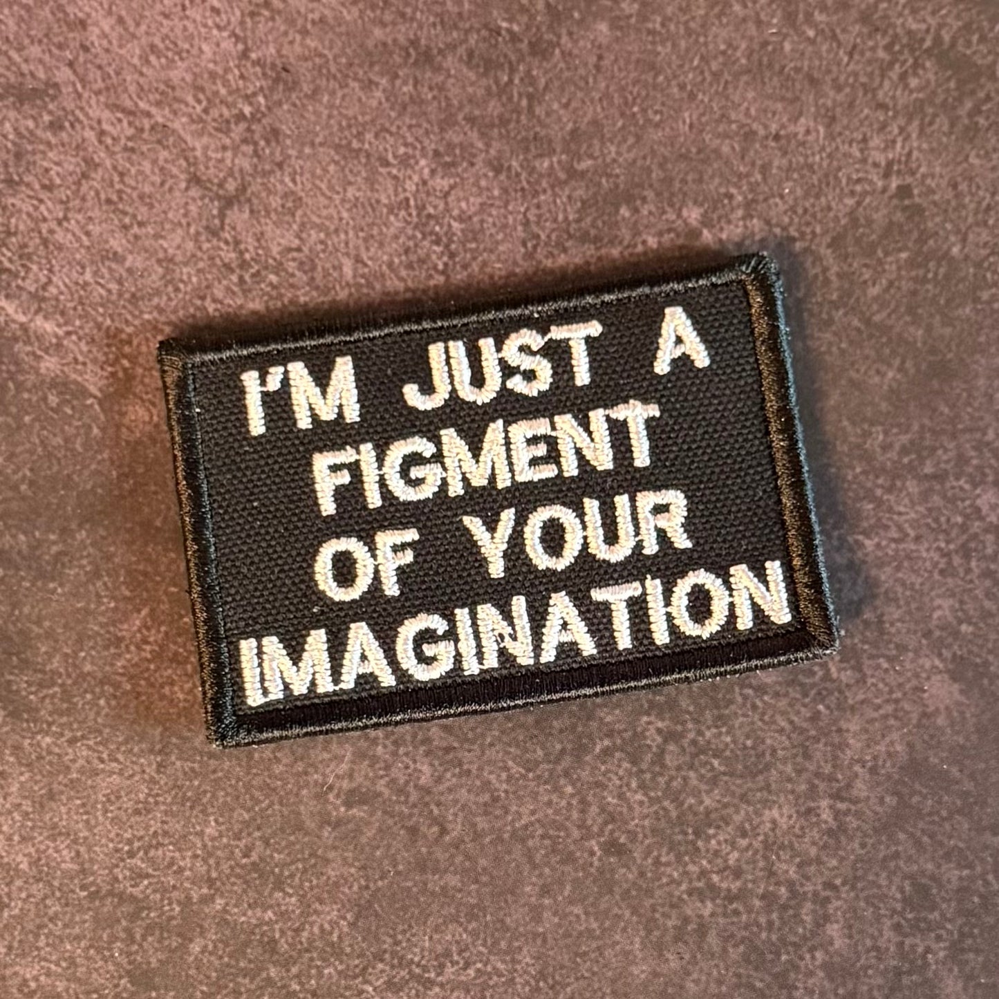 A rectangular embroidered patch with a black background and white text reading 'I'M JUST A FIGMENT OF YOUR IMAGINATION' on a brown textured fabric surface.