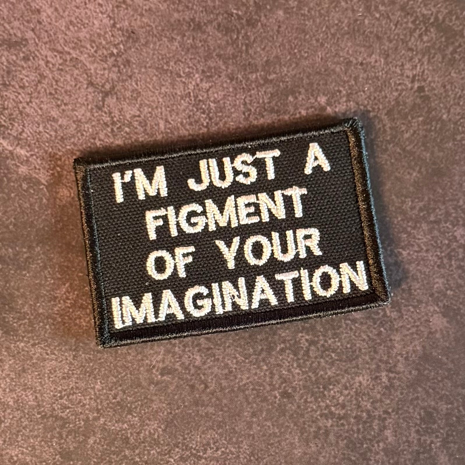A rectangular embroidered patch with a black background and white text reading 'I'M JUST A FIGMENT OF YOUR IMAGINATION' on a brown textured fabric surface.