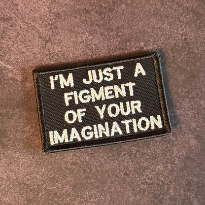 A rectangular embroidered patch with a black background and white text reading 'I'M JUST A FIGMENT OF YOUR IMAGINATION' on a brown textured fabric surface.