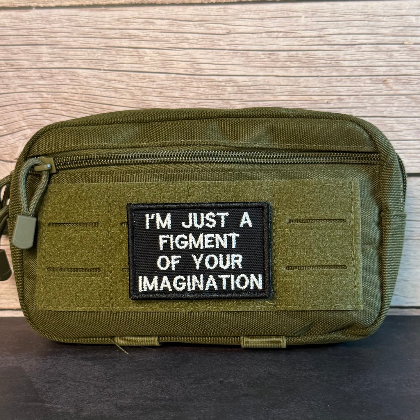 An olive green tactical pouch with a black rectangular patch that reads 'I'M JUST A FIGMENT OF YOUR IMAGINATION' in white text, set against a light wood background.
