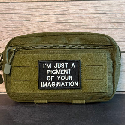An olive green tactical pouch with a black rectangular patch that reads 'I'M JUST A FIGMENT OF YOUR IMAGINATION' in white text, set against a light wood background.