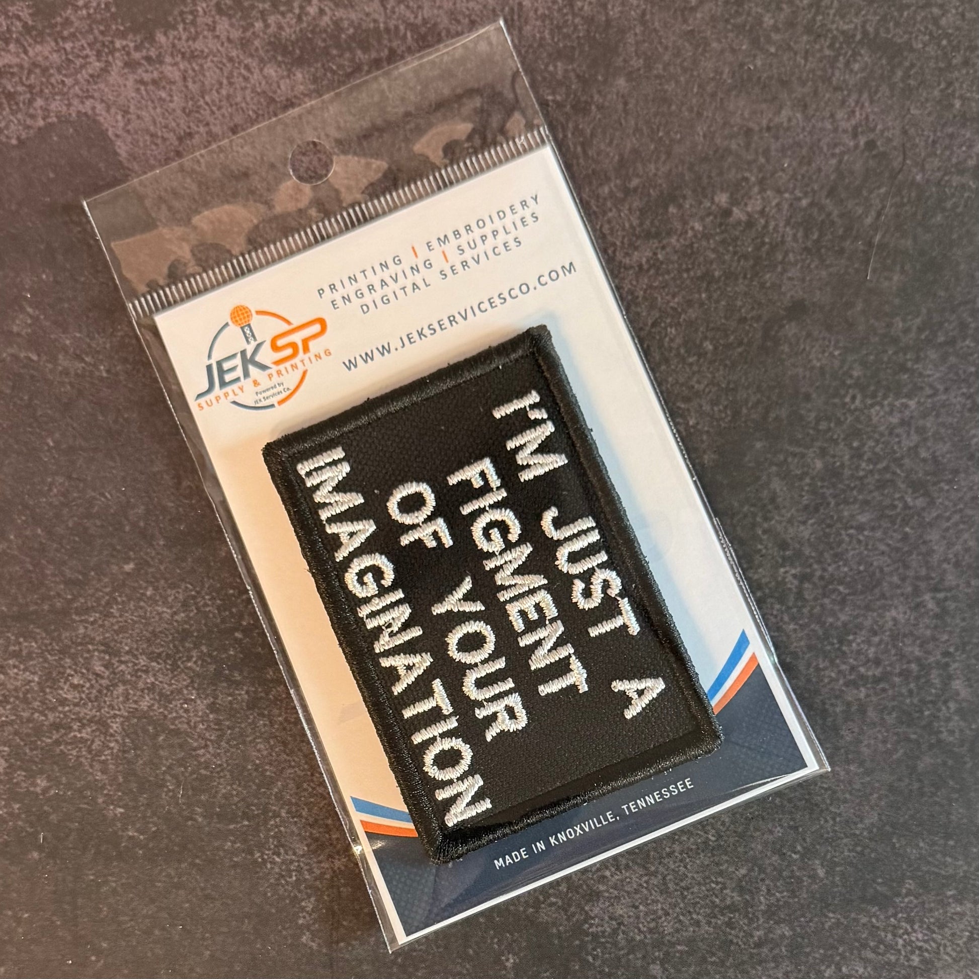 An embroidered black patch with white text reading 'I'M JUST A FIGMENT OF YOUR IMAGINATION' inside clear retail packaging on a dark textured surface.