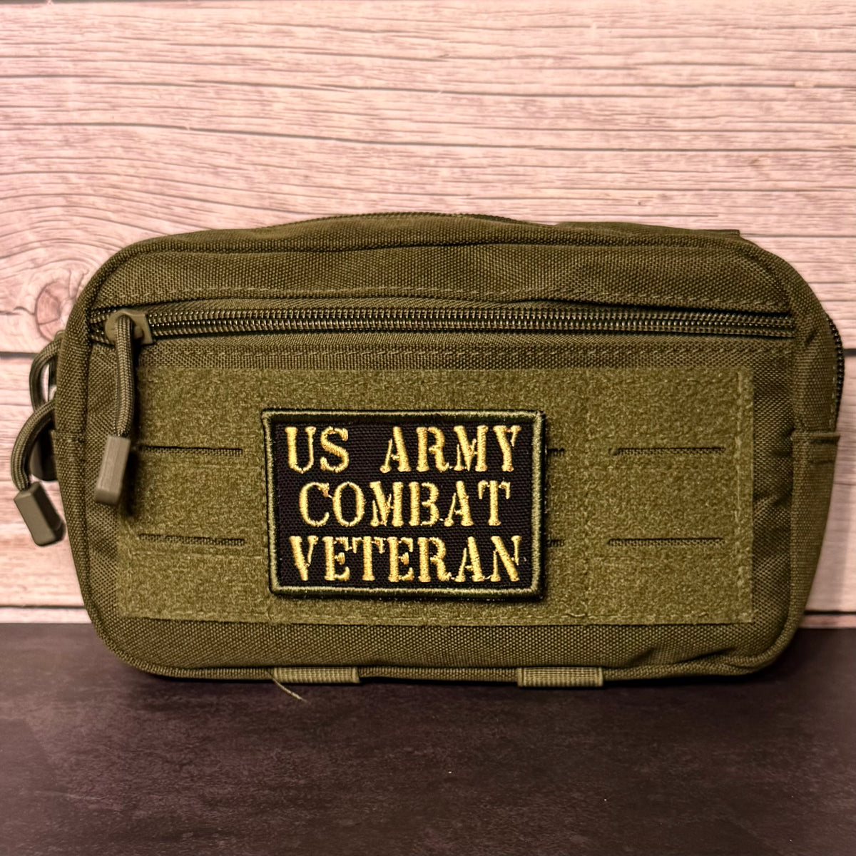An olive green tactical pouch with a front zipper and a black rectangular patch reading 'US ARMY COMBAT VETERAN' in yellow lettering, shown against a wood plank background.