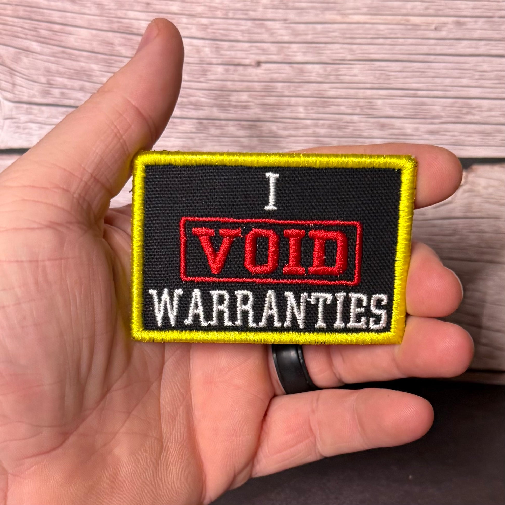 A hand holding a rectangular embroidered patch that reads 'I VOID WARRANTIES' in red and white text on a black background with a yellow border, against a wood plank background.