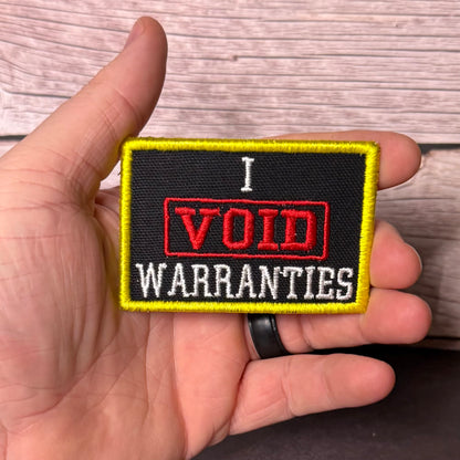 A hand holding a rectangular embroidered patch that reads 'I VOID WARRANTIES' in red and white text on a black background with a yellow border, against a wood plank background.