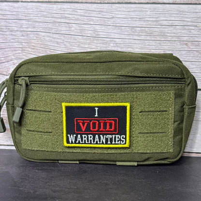 An olive green tactical pouch with a front zipper and a rectangular embroidered patch that reads 'I VOID WARRANTIES' in red and white on a black background with a yellow border, set against a gray wood-plank backdrop.