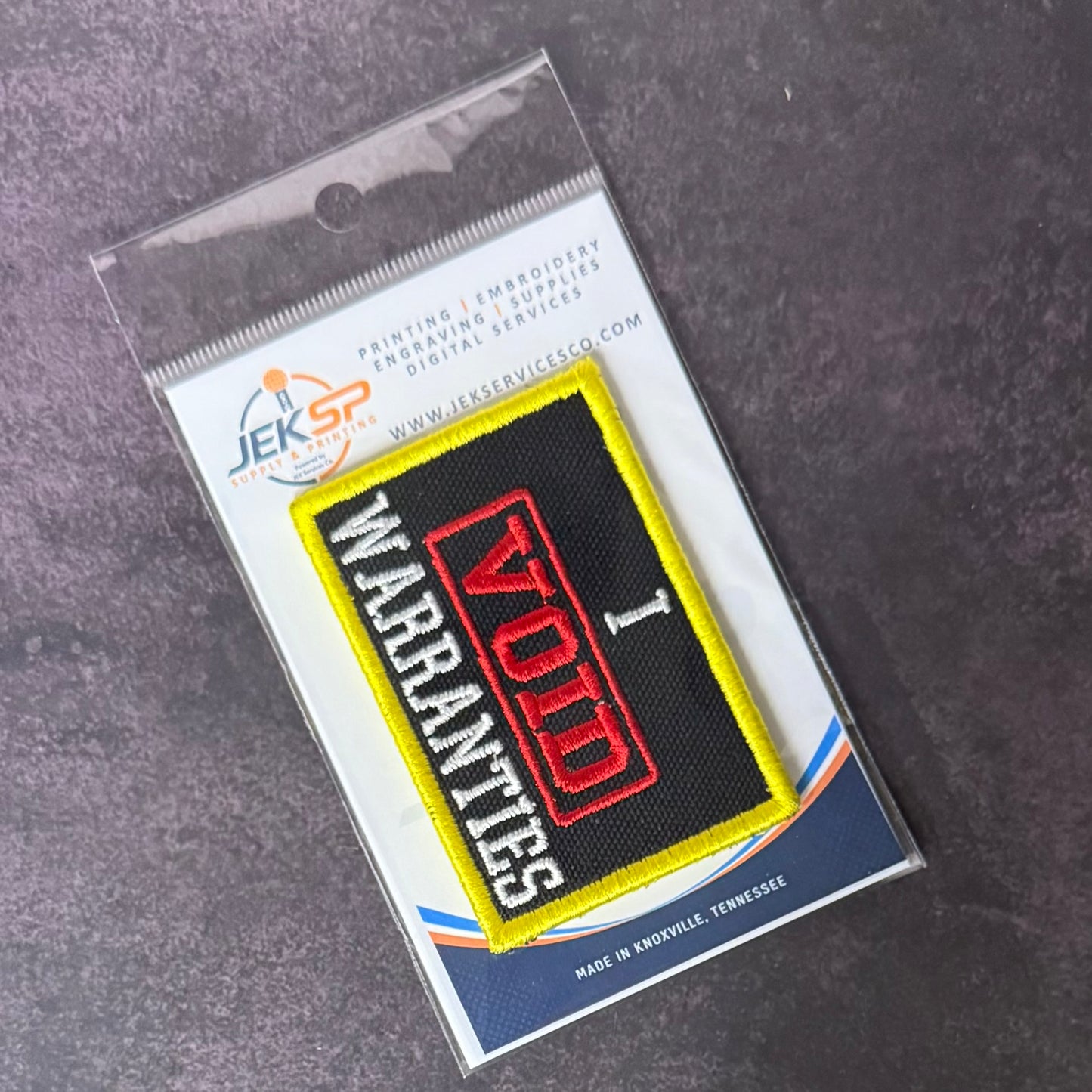 An embroidered patch reading 'WARRANTIES VOID' in red, white, and black with a yellow border, sealed in a clear plastic package on a branded card over a dark textured surface.