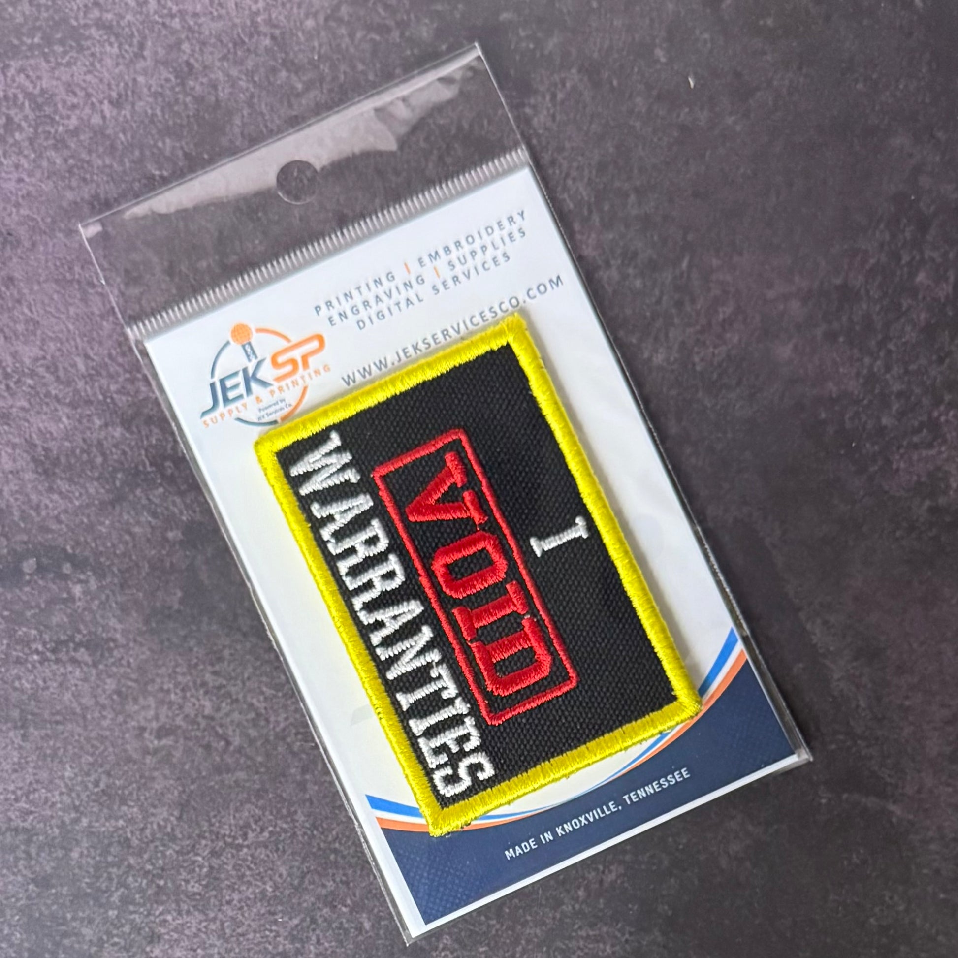 An embroidered patch reading 'WARRANTIES VOID' in red, white, and black with a yellow border, sealed in a clear plastic package on a branded card over a dark textured surface.