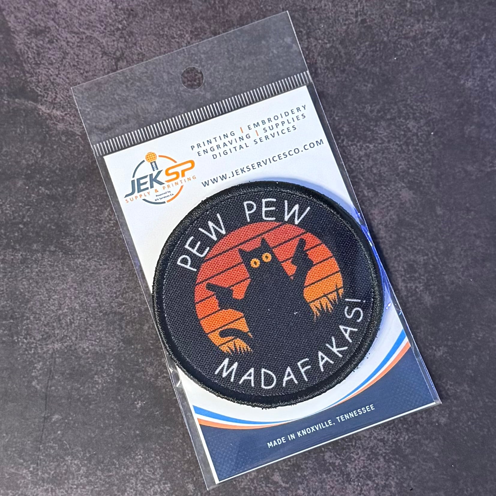 A circular embroidered patch on a printed backing card, photographed on a dark textured surface. The patch reads 'PEW PEW MADAFAKASI' around a black cat silhouette against an orange sunset.
