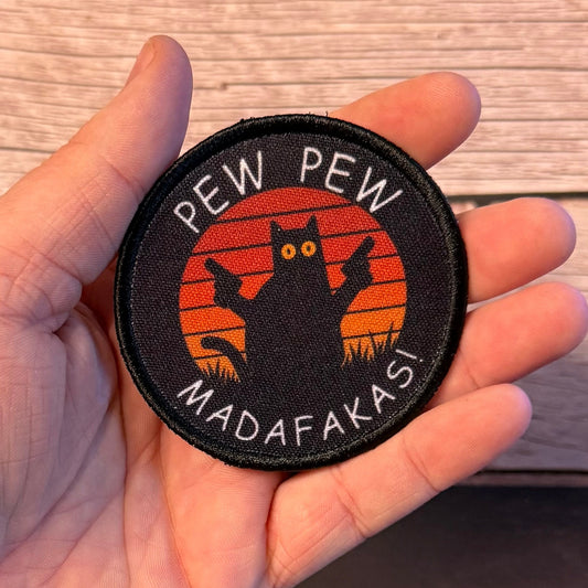 A circular embroidered patch held in a hand against a light wood plank background. The patch has a black border and background with a black cat silhouette in front of orange sunset-style stripes, plus white text reading 'PEW PEW MADAFAKASI'.