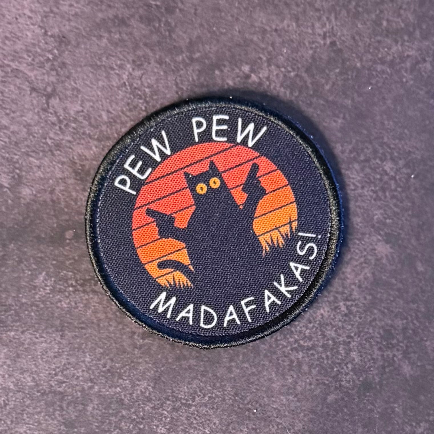 A circular embroidered patch featuring a black cat silhouette in front of an orange sunset, with the words 'PEW PEW' at the top and 'MADAFAKASI' at the bottom, placed on a textured gray surface.