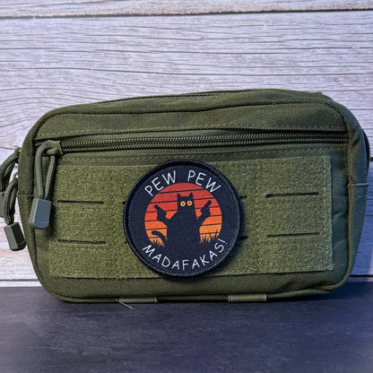 An olive green tactical pouch with a circular embroidered patch featuring a black cat silhouette against an orange sunset and the text 'PEW PEW MADAFAKASI'.