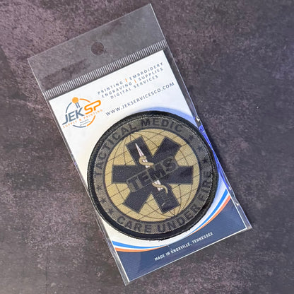 A circular tactical medic patch with a black Star of Life and an ambulance symbol on a tan background, sealed in a clear plastic package with a white and blue JEKSP card backing, photographed on a dark textured surface.