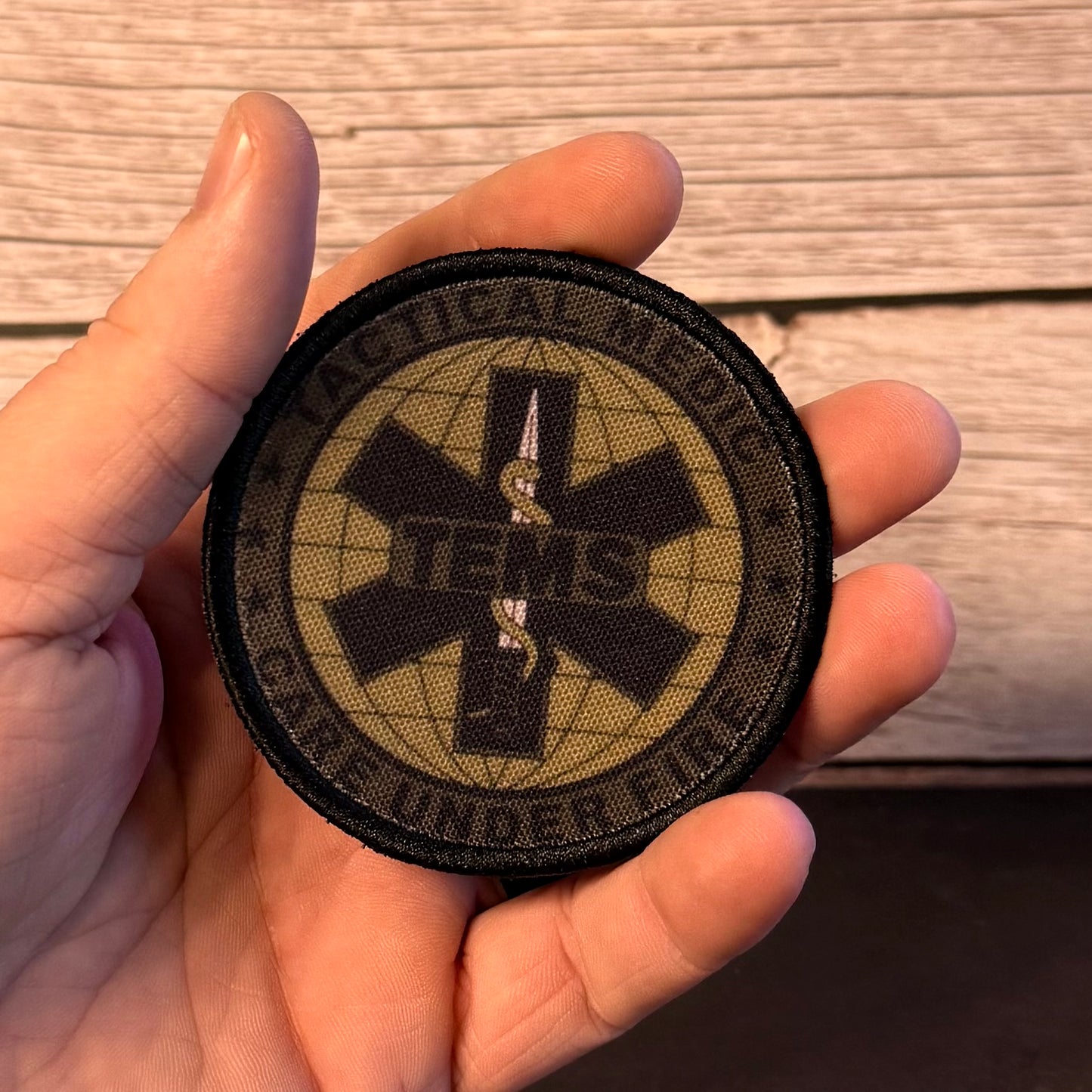A hand holding a round tactical medic patch with a black border, featuring a black cross and snake symbol over a tan grid background with the text 'VITALIC MEDIC' and 'ONE UNDER FIRE'.