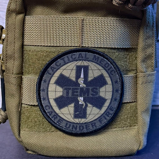 A close-up of a tactical pouch in olive drab nylon with a circular embroidered patch featuring a dark blue Star of Life and the text 'Tactical Medic Care Under Fire' and 'ITEMS'.