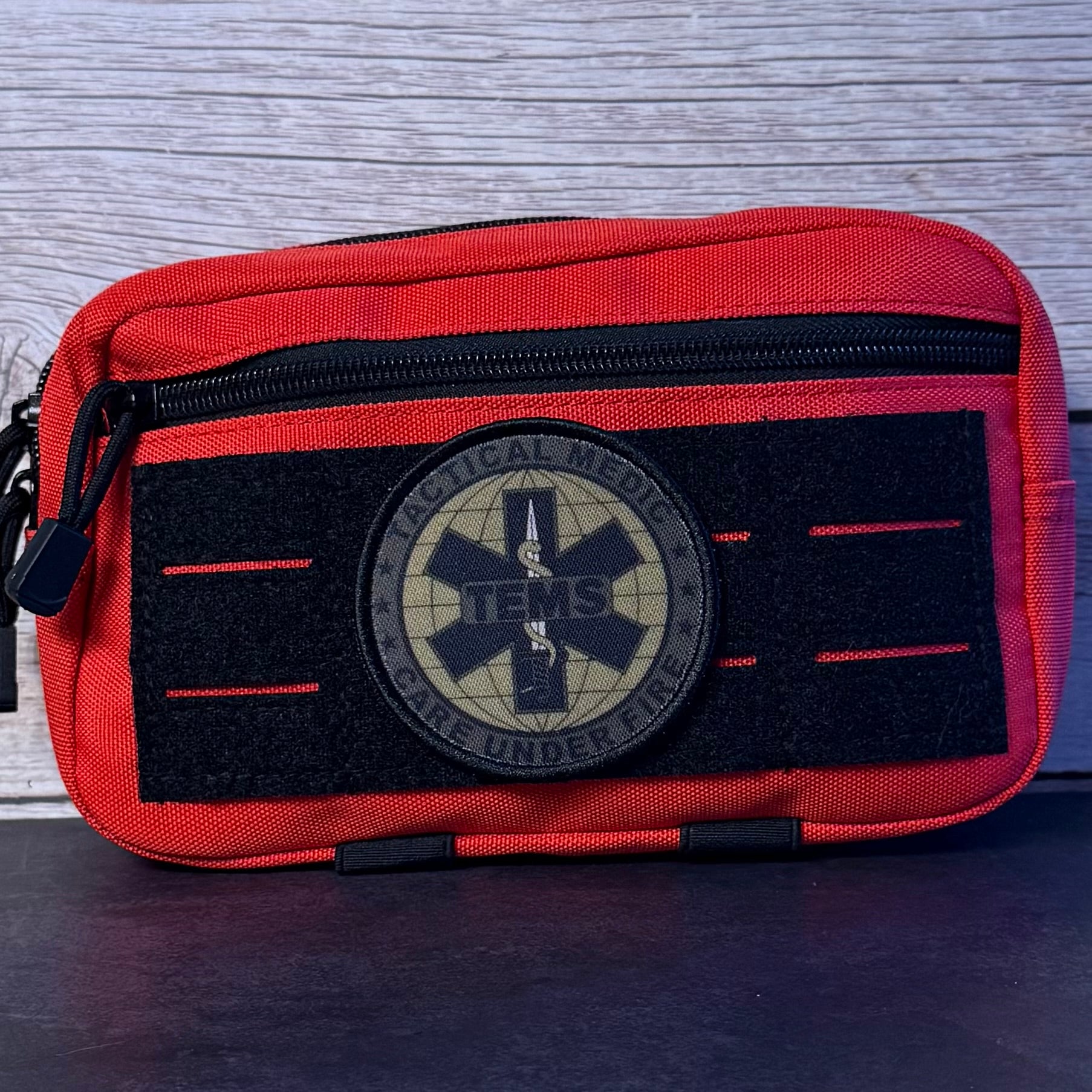 A tactical medical pouch in red and black nylon, featuring a circular 'Tactical Medical TENS Care Under Fire' patch on the front, set against a gray wood-grain background.