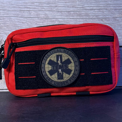 A tactical medical pouch in red and black nylon, featuring a circular 'Tactical Medical TENS Care Under Fire' patch on the front, set against a gray wood-grain background.