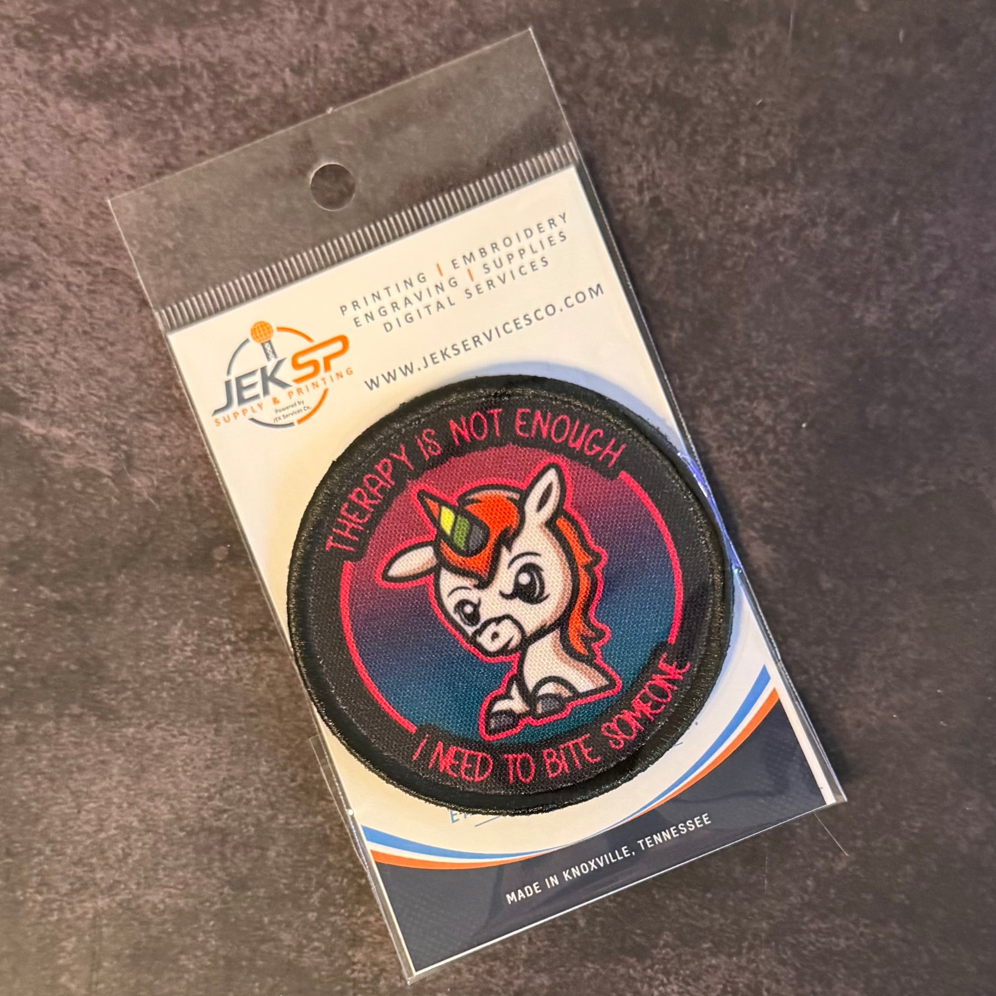 A circular embroidered patch featuring a cartoon unicorn with a horn and the text 'THERAPY IS NOT ENOUGH I NEED TO BITE SOMEONE', attached to a printed backing card from JEKSP Service Co. The card is photographed on a textured gray surface.