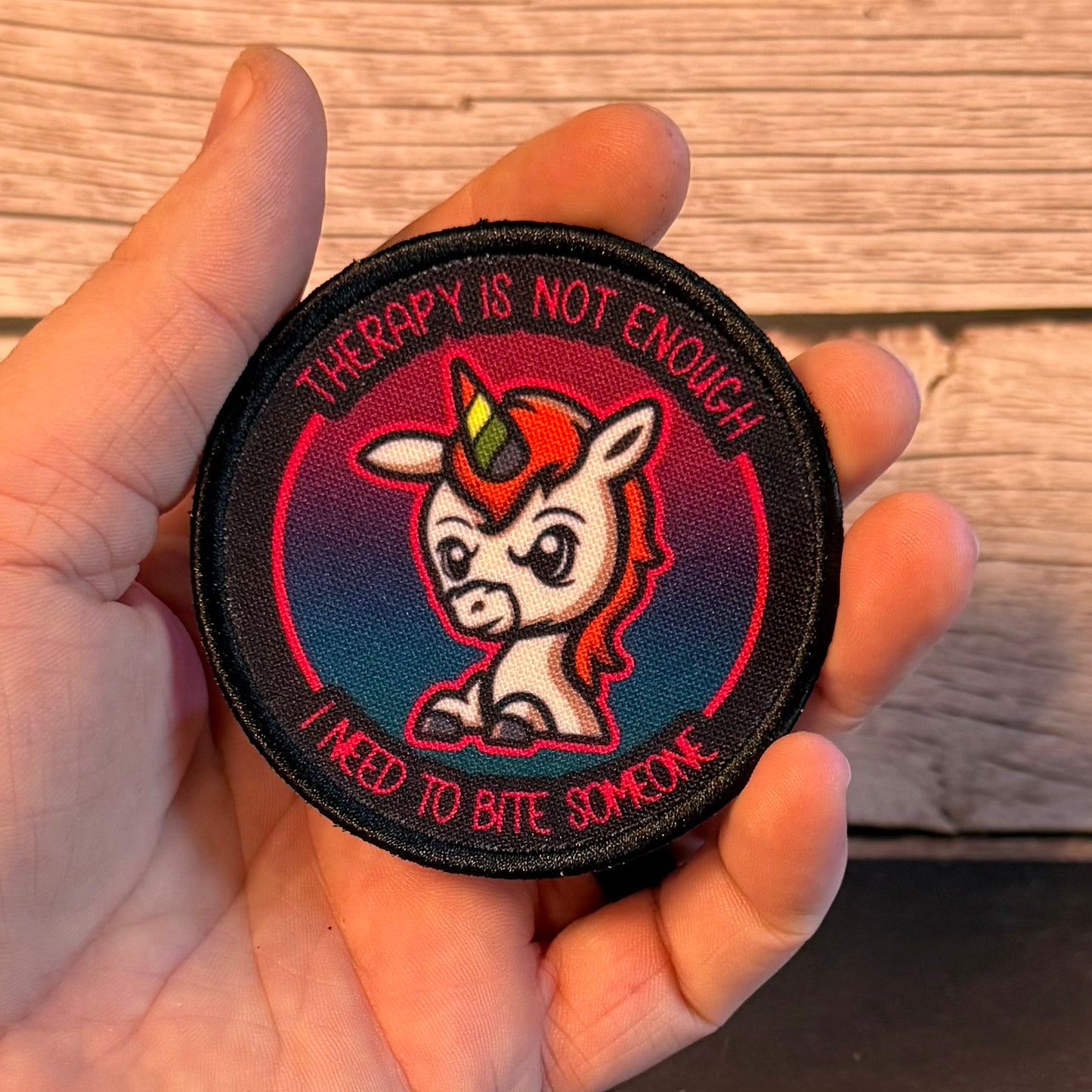 A hand holding a round embroidered patch featuring a cartoon unicorn with a red horn and the text 'THERAPY IS NOT ENOUGH I NEED TO BITE SOMEONE' against a wood plank background.