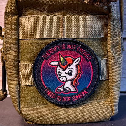 A circular embroidered patch featuring a cartoon unicorn with the text 'THERAPY IS NOT ENOUGH' and 'I NEED TO BITE SOMEONE', attached to a tan tactical pouch.