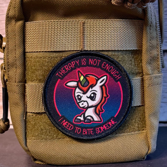 A circular embroidered patch featuring a cartoon unicorn with the text 'THERAPY IS NOT ENOUGH' and 'I NEED TO BITE SOMEONE', attached to a tan tactical pouch.