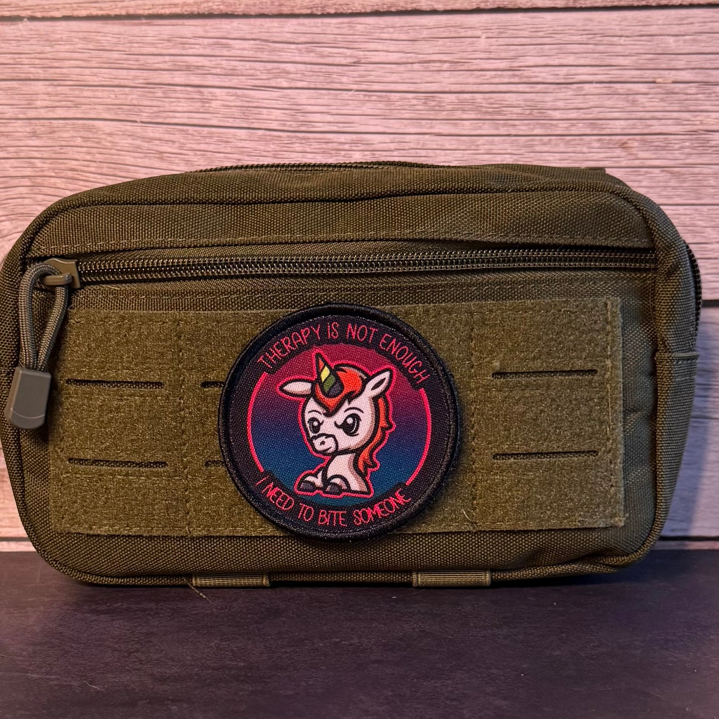 An olive green tactical pouch with a circular embroidered patch featuring a cartoon unicorn and the text 'THERAPY IS NOT ENOUGH! I NEED TO BITE SOMEONE'.