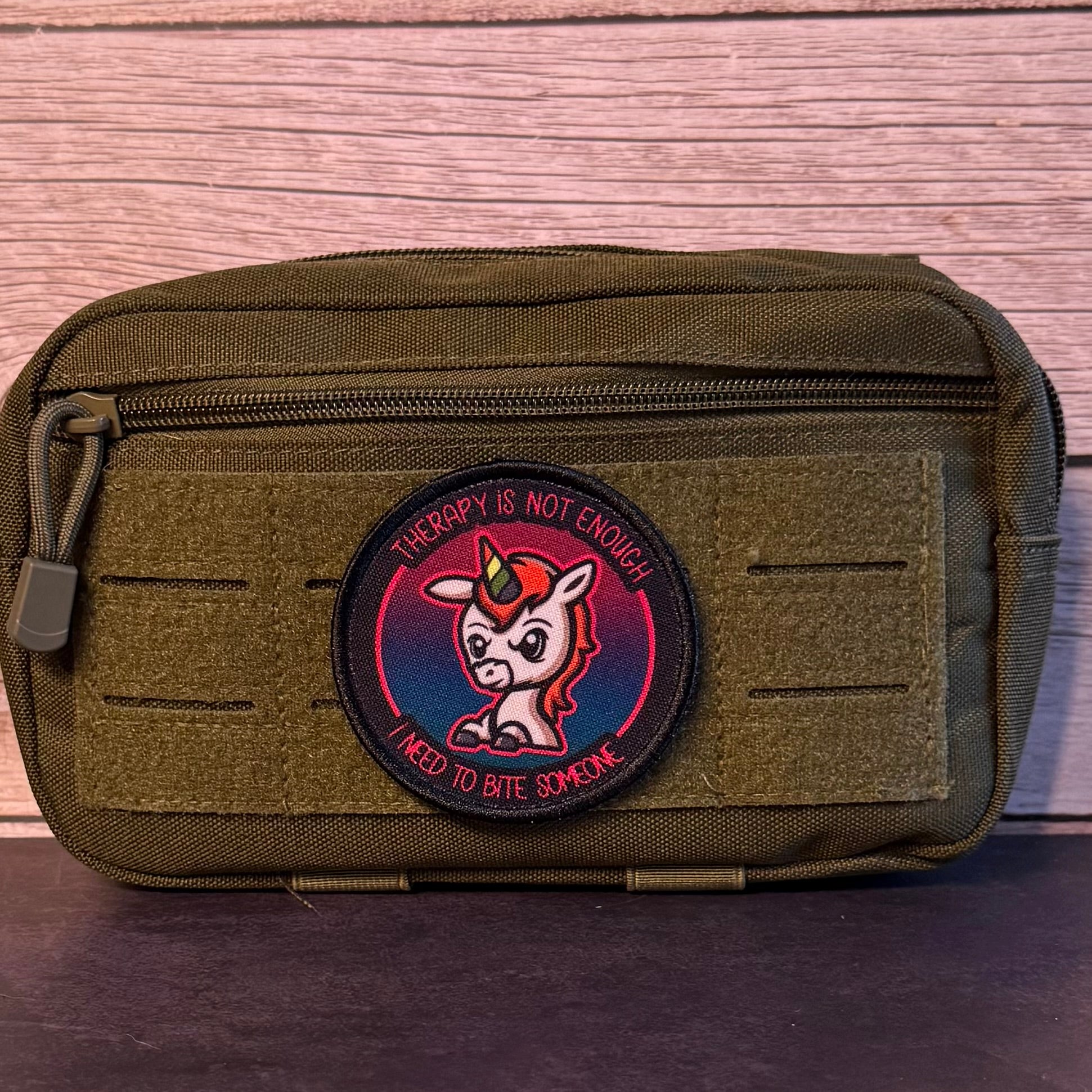 An olive green tactical pouch with a circular embroidered patch featuring a cartoon unicorn and the text 'THERAPY IS NOT ENOUGH! I NEED TO BITE SOMEONE'.