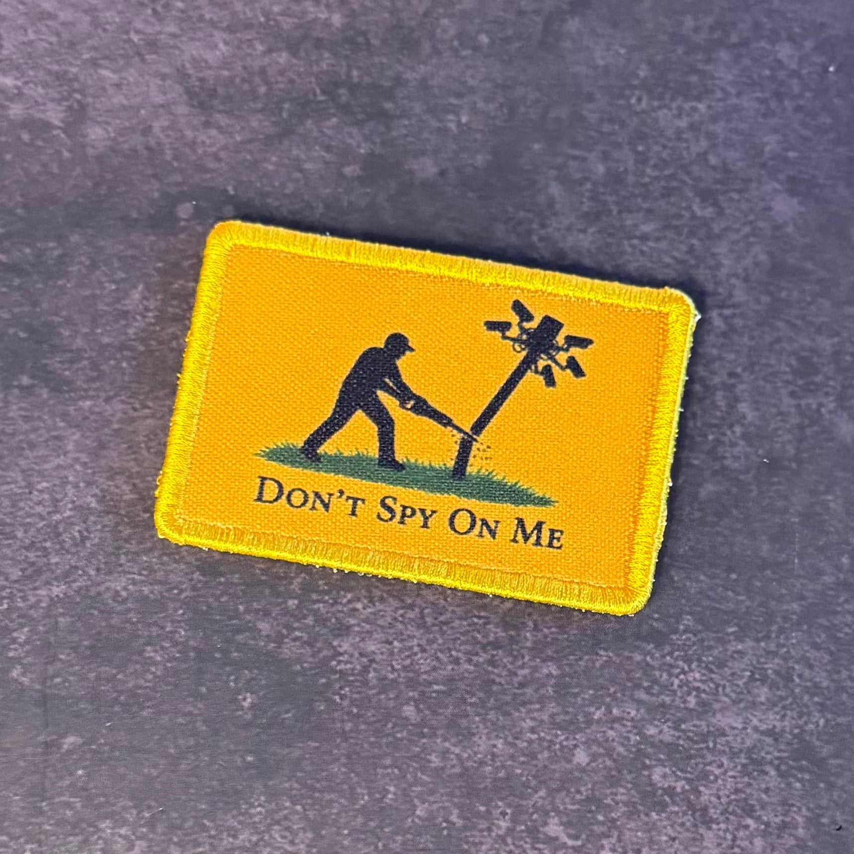 A rectangular yellow embroidered patch with a black silhouette of a person cutting a wire pole, with the text 'DON'T SPY ON ME' below, placed on a textured dark gray surface.