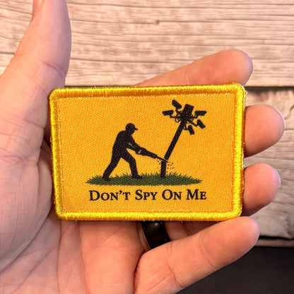 A hand holding a rectangular yellow embroidered patch with a black silhouette of a person cutting a wire pole and the text 'DON'T SPY ON ME' against a wood plank background.