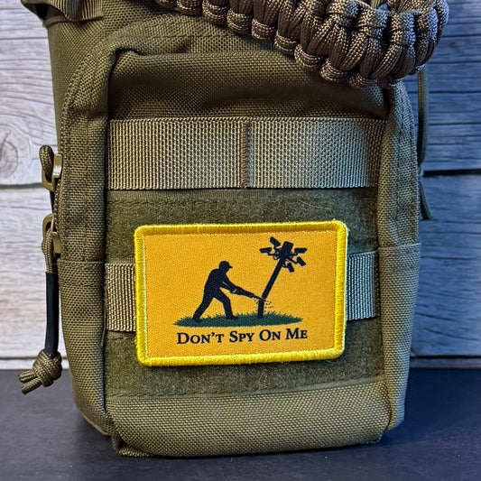 An olive green tactical pouch with a yellow embroidered patch that reads 'DON'T SPY ON ME' and shows a black silhouette of a person hanging from a tree. The pouch has a paracord strap and is photographed against a dark wood background.