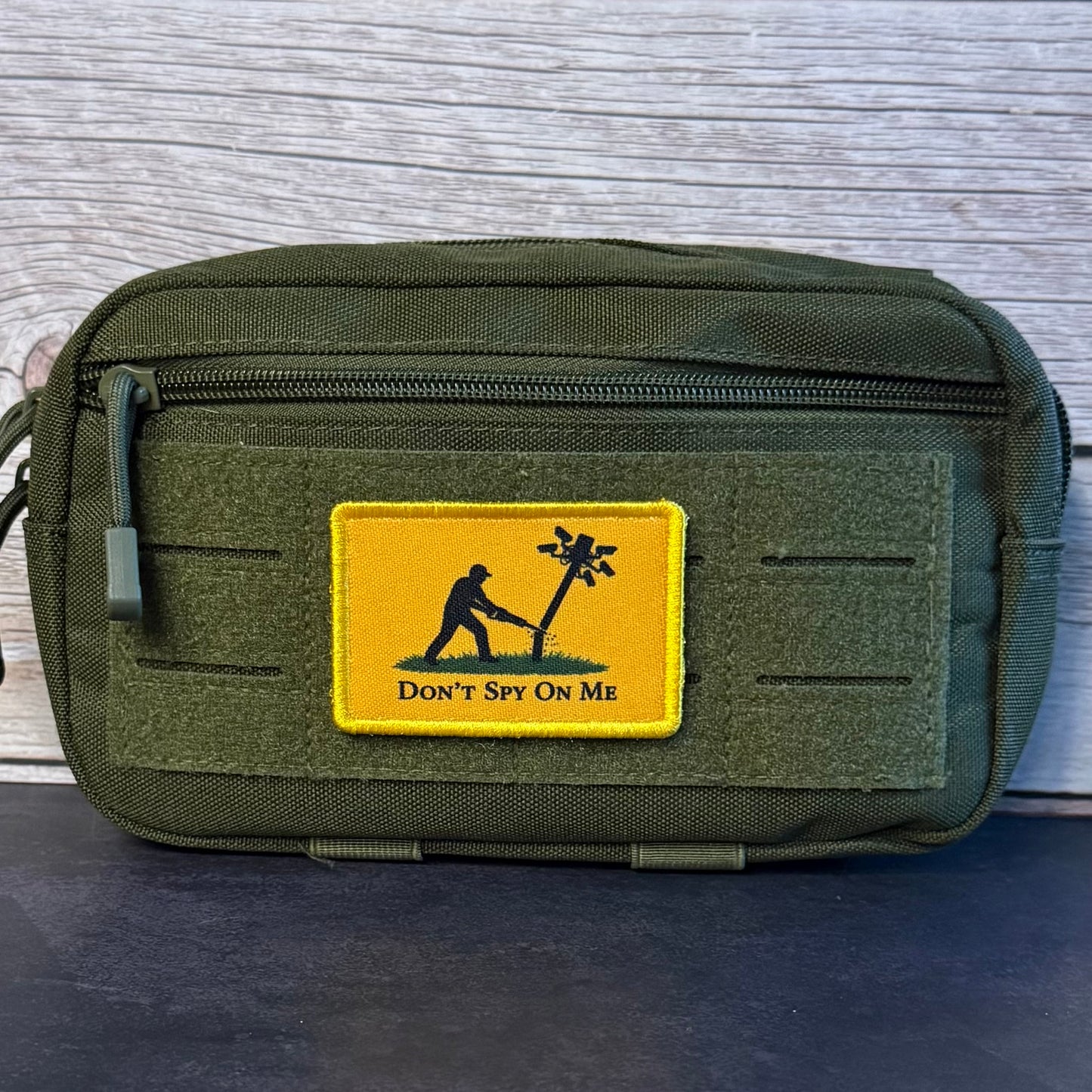 An olive green tactical pouch with a front zipper and a yellow rectangular patch featuring a black silhouette of a person planting a palm tree and the text 'DON'T SPY ON ME'. The pouch is photographed against a light gray wood plank background.