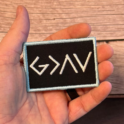 A rectangular embroidered patch held in a hand, featuring white embroidered runes on a black background with a light blue border, set against a wooden surface.