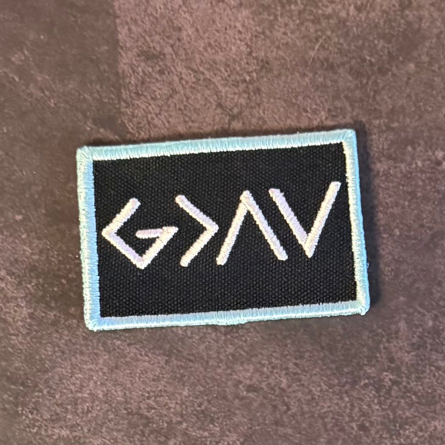 A rectangular embroidered patch with a black center, white stylized lettering, and a light blue border, placed on a brown textured surface.