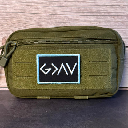 An olive green tactical pouch with a black rectangular patch featuring the white embroidered letters 'GNAV' and a light blue border, set against a rustic wood background.