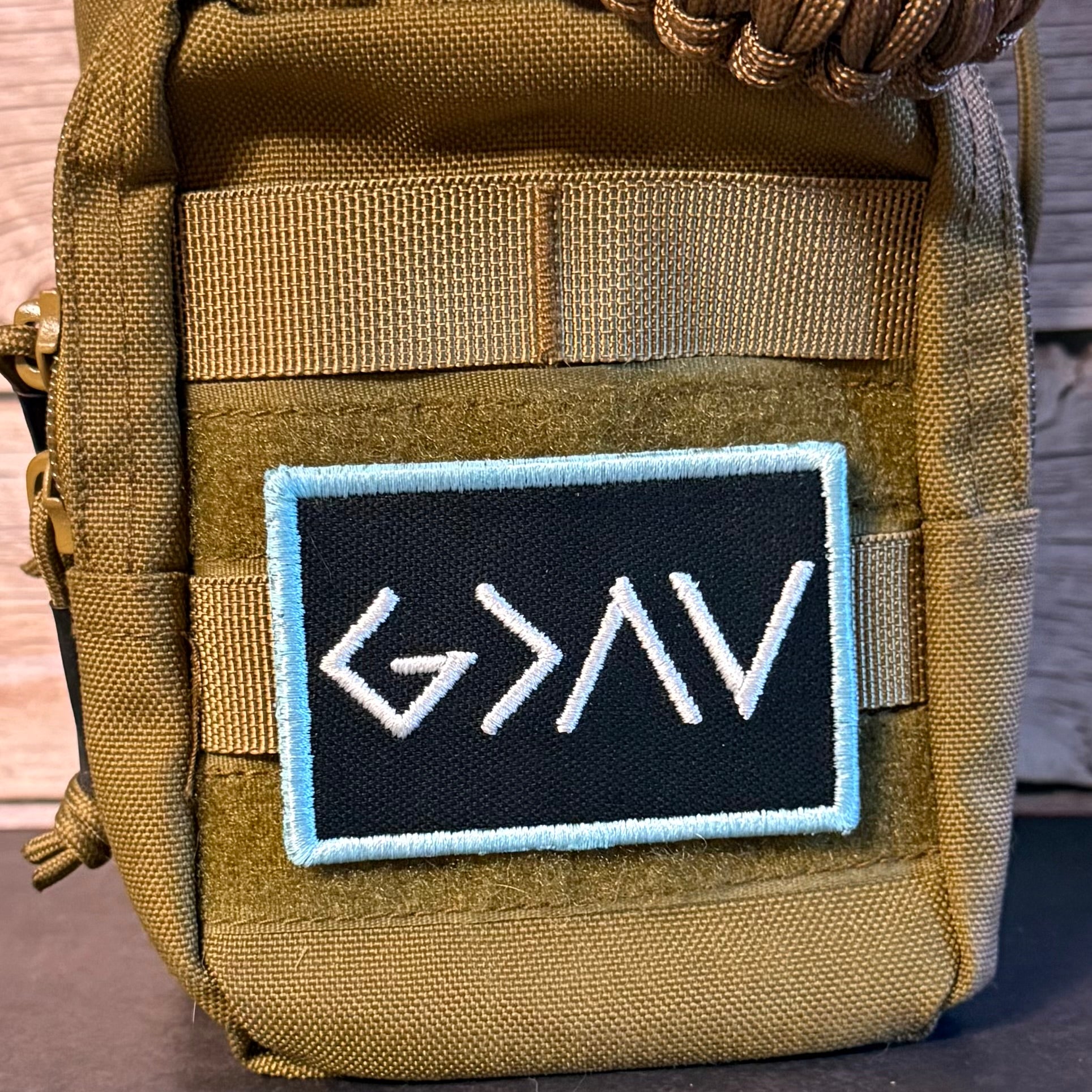 Close-up of a tactical pouch in olive drab green with a black rectangular embroidered patch featuring white stylized lettering and a light blue border.