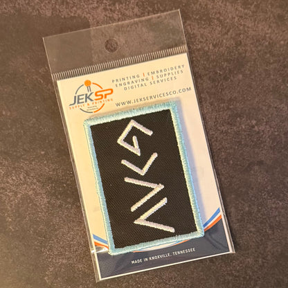 A black rectangular embroidered patch with white stylized lettering and a light blue border, sealed in a clear plastic retail package with a branded card backing. The visible brand on the packaging is JEKSP Supply Co.